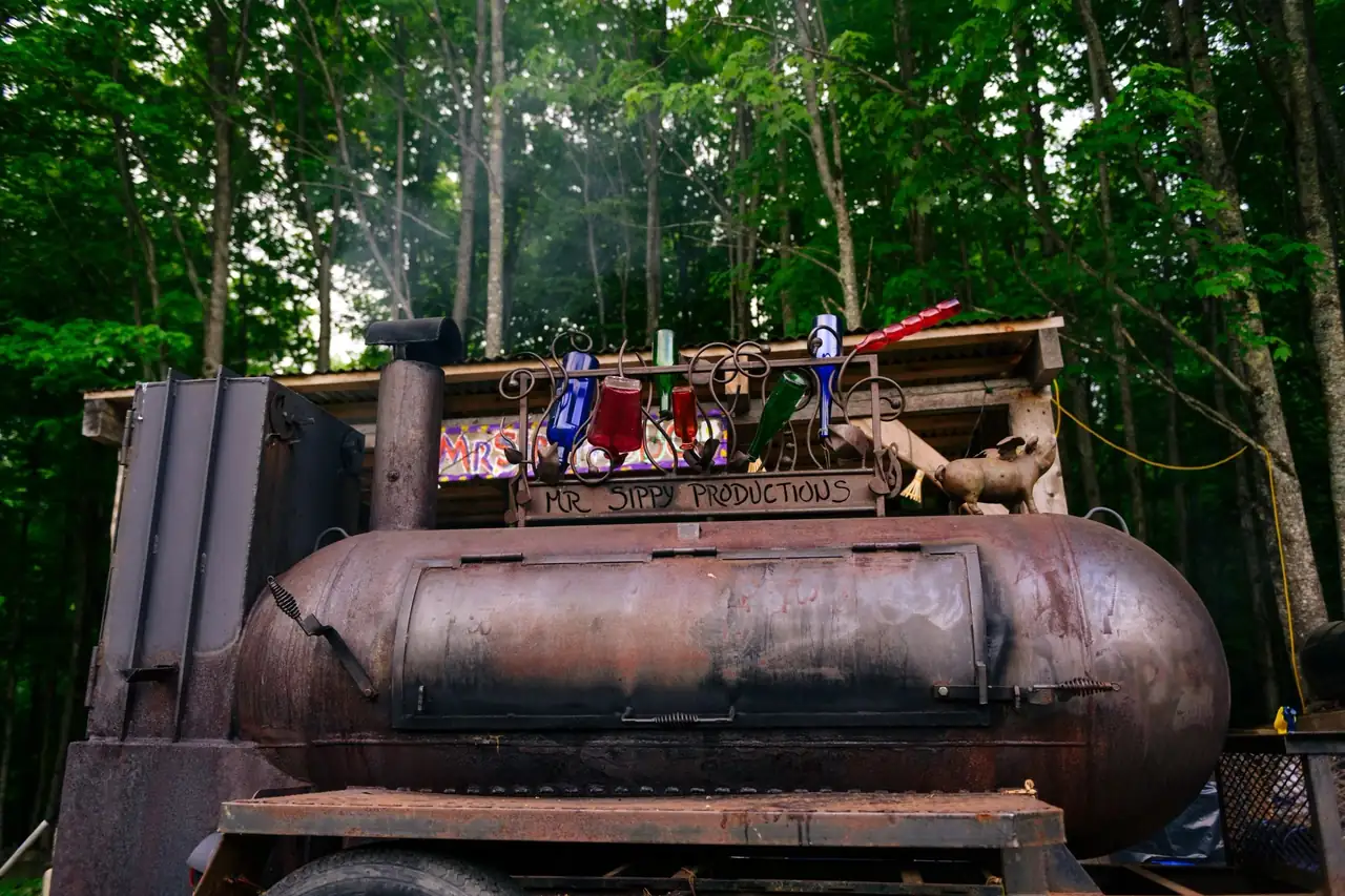MrSippy's Lang reverse-flow smoker sending up hickory smoke in the New Hampshire woods, crowned by the Mr Sippy Productions bottle tree