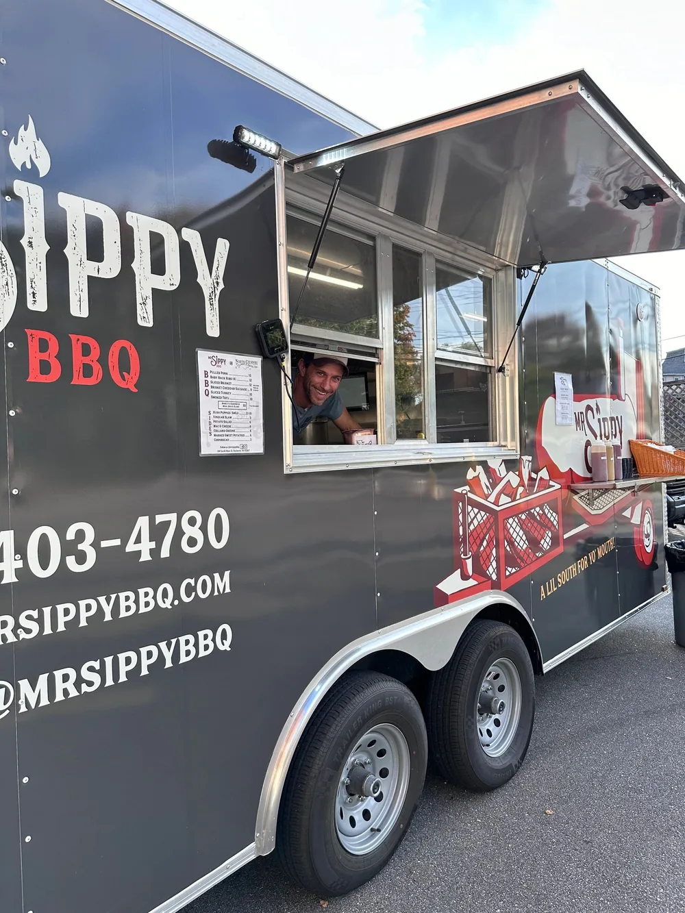 The black 18-foot MrSippy BBQ trailer with the serving window open and an order being handed out