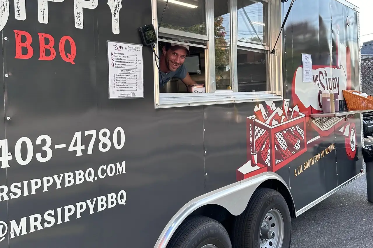 The MrSippy BBQ trailer at an event, serving window open, an order being handed to a guest
